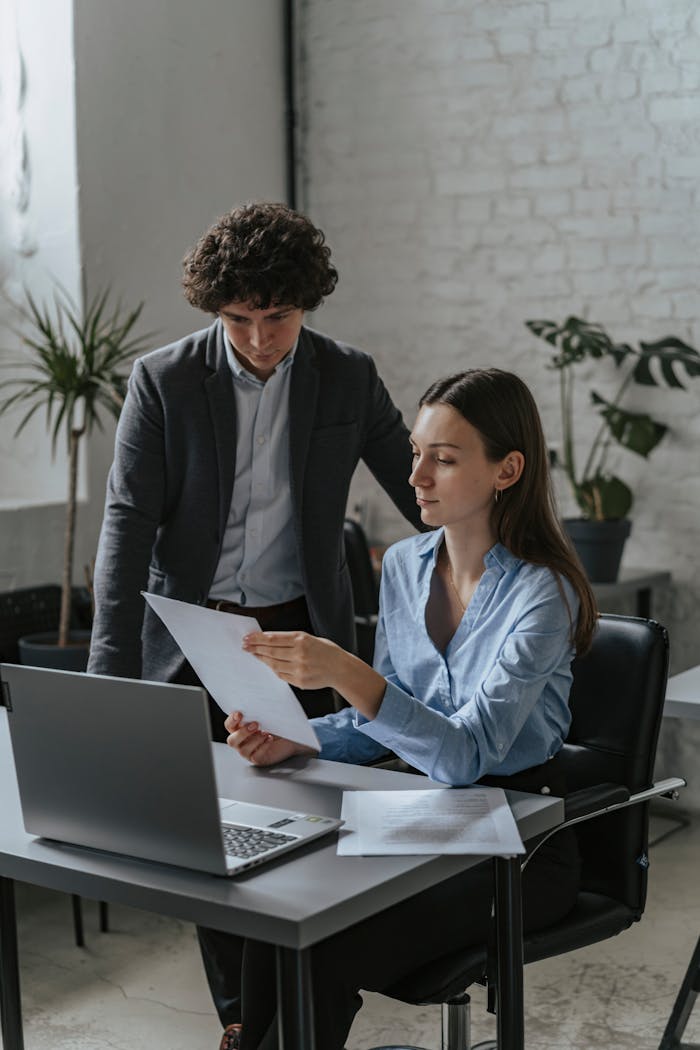 Two professionals in an office setting reviewing documents on a laptop.